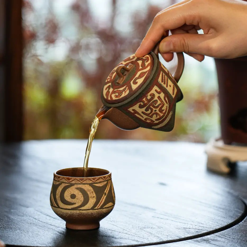 Tea gracefully pouring from the spout of the Cloud & Thunder Yixing teapot into a matching patterned teacup during a mindful Gongfu tea session.