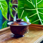 Decorative but functional gaiwan on a wooden surface with green foliage in the background
