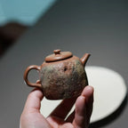 Close-up lifestyle shot of a hand holding the Desert Mirage Yixing teapot over a white tea boat, emphasizaing its tough, rugged surface and the vibrant, free-style hand-painted colored clay accents.
