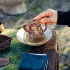 Close-up lifestyle shot of a hand holding the Formless Zen Yixing teapot over a traditional tea boat on an outdoor wooden table, highlighting the vivid, hand-painted Dunhuang Buddhist statues.