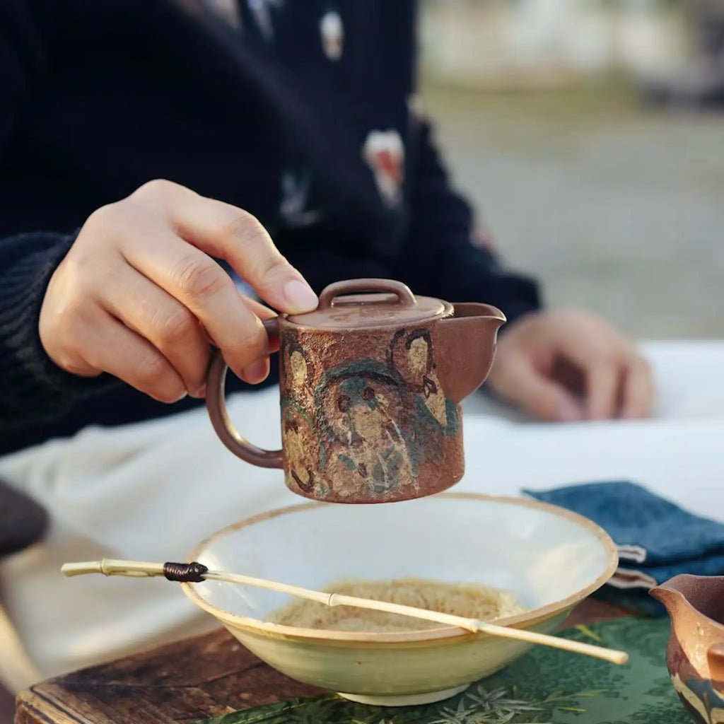 Lifestyle view of a hand holding the Formless Zen Yixing teapot outdoors, revealing the reverse side of the exquisite hand-painted Dunhuang Buddhist artwork resting above a wooden tea table and tea boat.