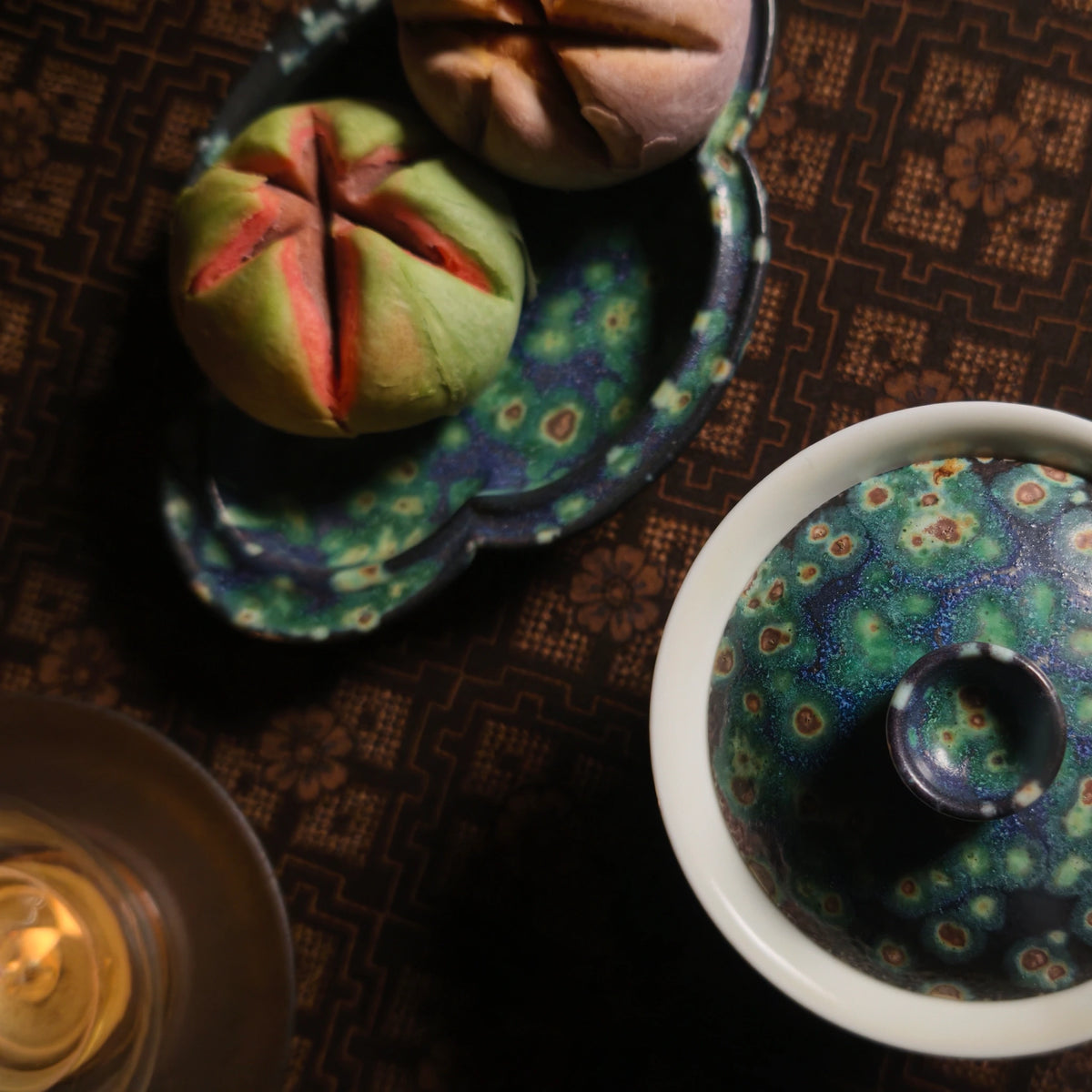 A close-up, top-down view of a ceramic tea set with a matching lidded cup and saucer, decorated with a vibrant blue, green, and purple floral pattern. A small candle is lit in the foreground on a dark brown patterned tablecloth.