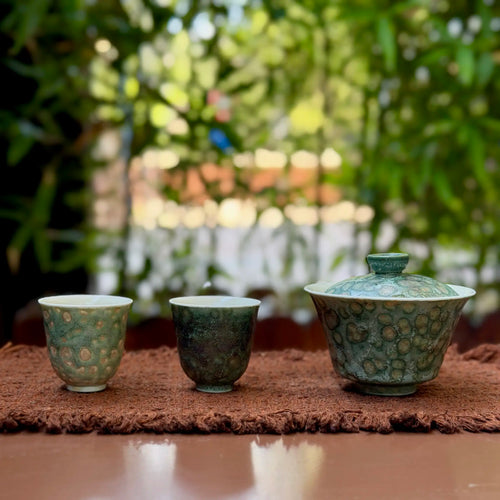 One porcelain gaiwan pairs with two matching teacups on a brown tea mat against a blurred green outdoor background