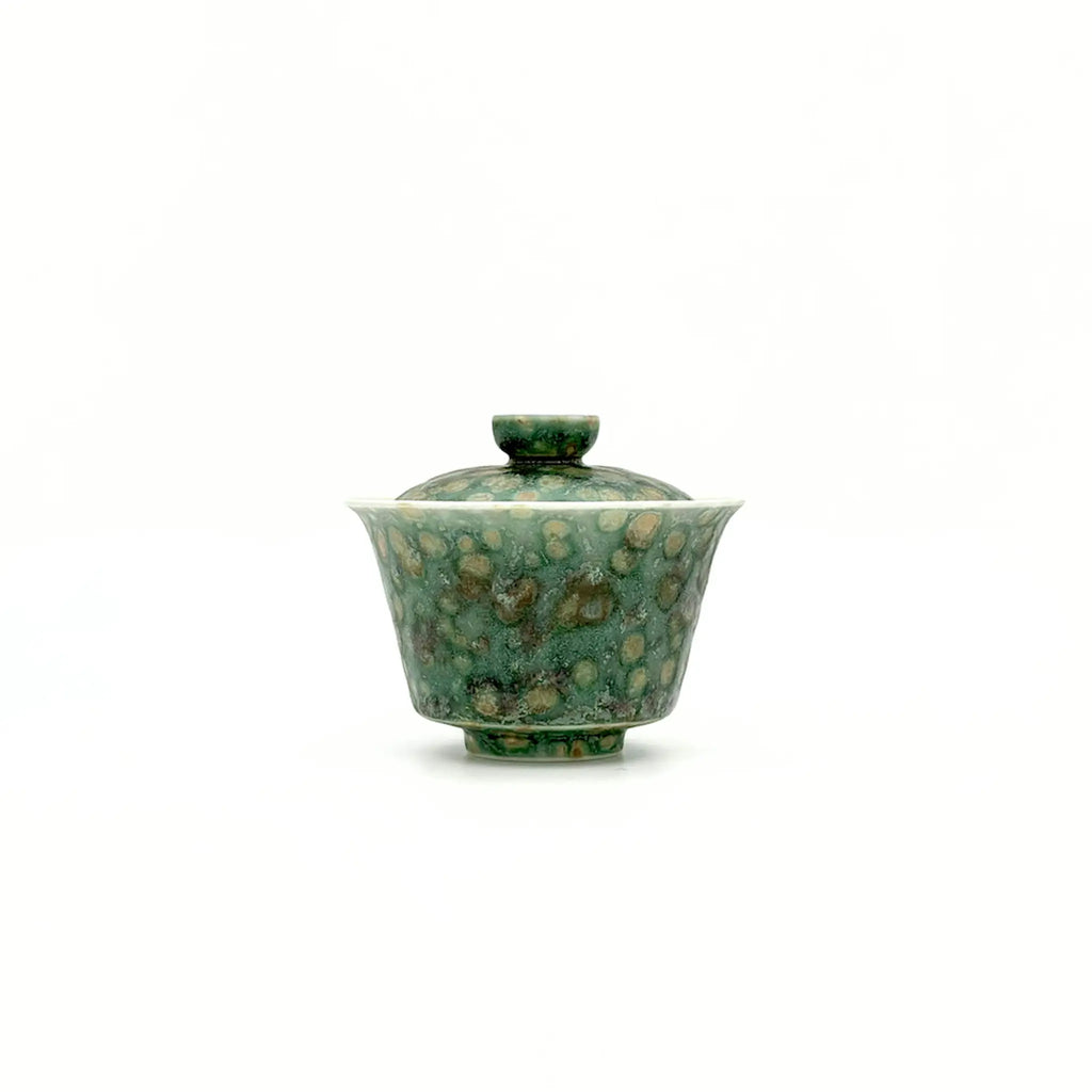A small glazed ceramic gaiwan or lidded teabowl with a mottled green and gold pattern, centered on a plain white background.