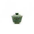 A small glazed ceramic gaiwan or lidded teabowl with a mottled green and gold pattern, centered on a plain white background.