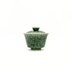 A small glazed ceramic gaiwan or lidded teabowl with a mottled green and gold pattern, centered on a plain white background.