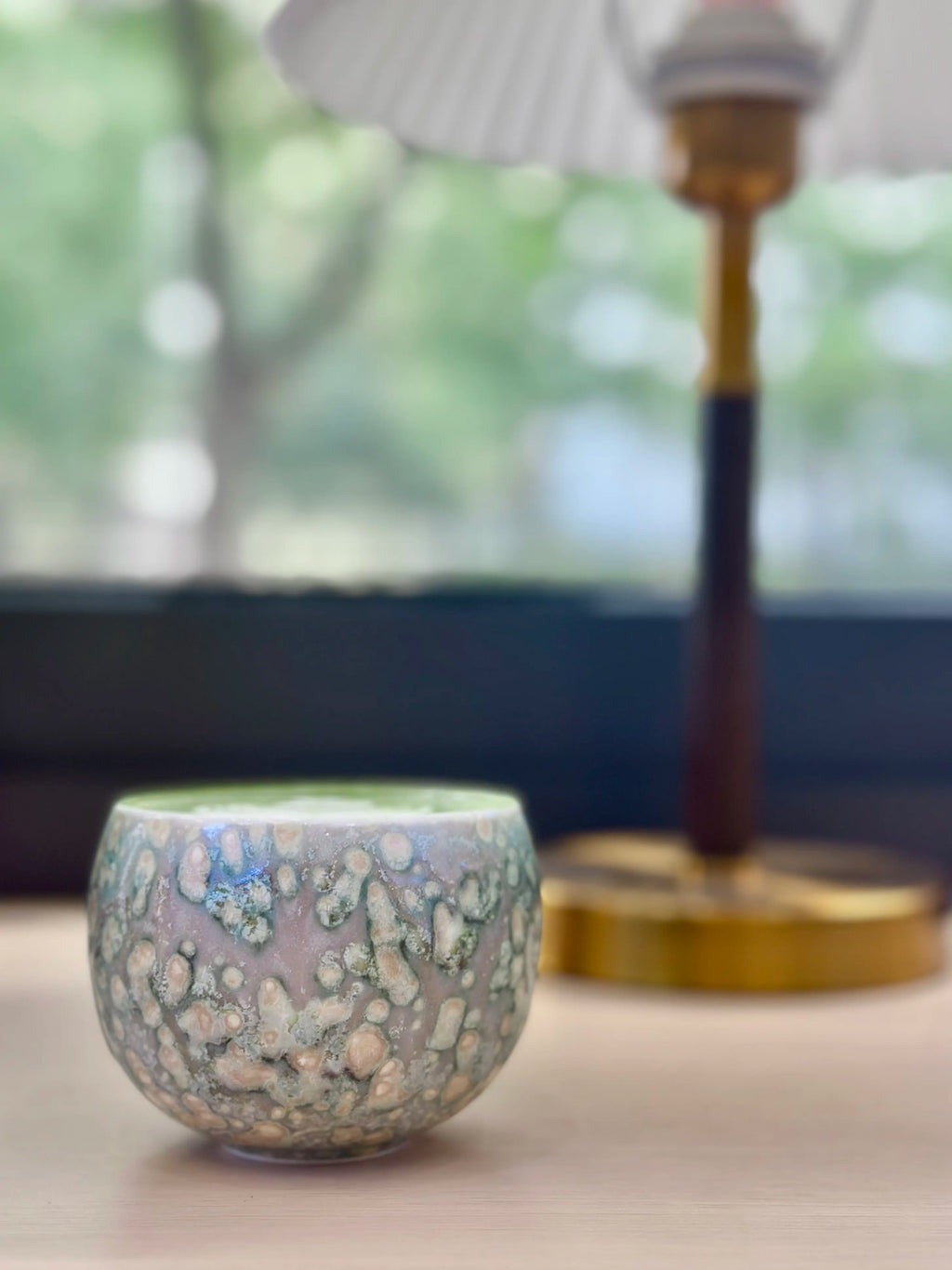 Lifestyle desk shot featuring the Glazara Emerald Current Matcha Latte Cup filled with fresh matcha. Backlit by a warm desk lamp against a window background, identifying the deep, jewel-toned green glaze and creating a cozy workspace atmosphere.