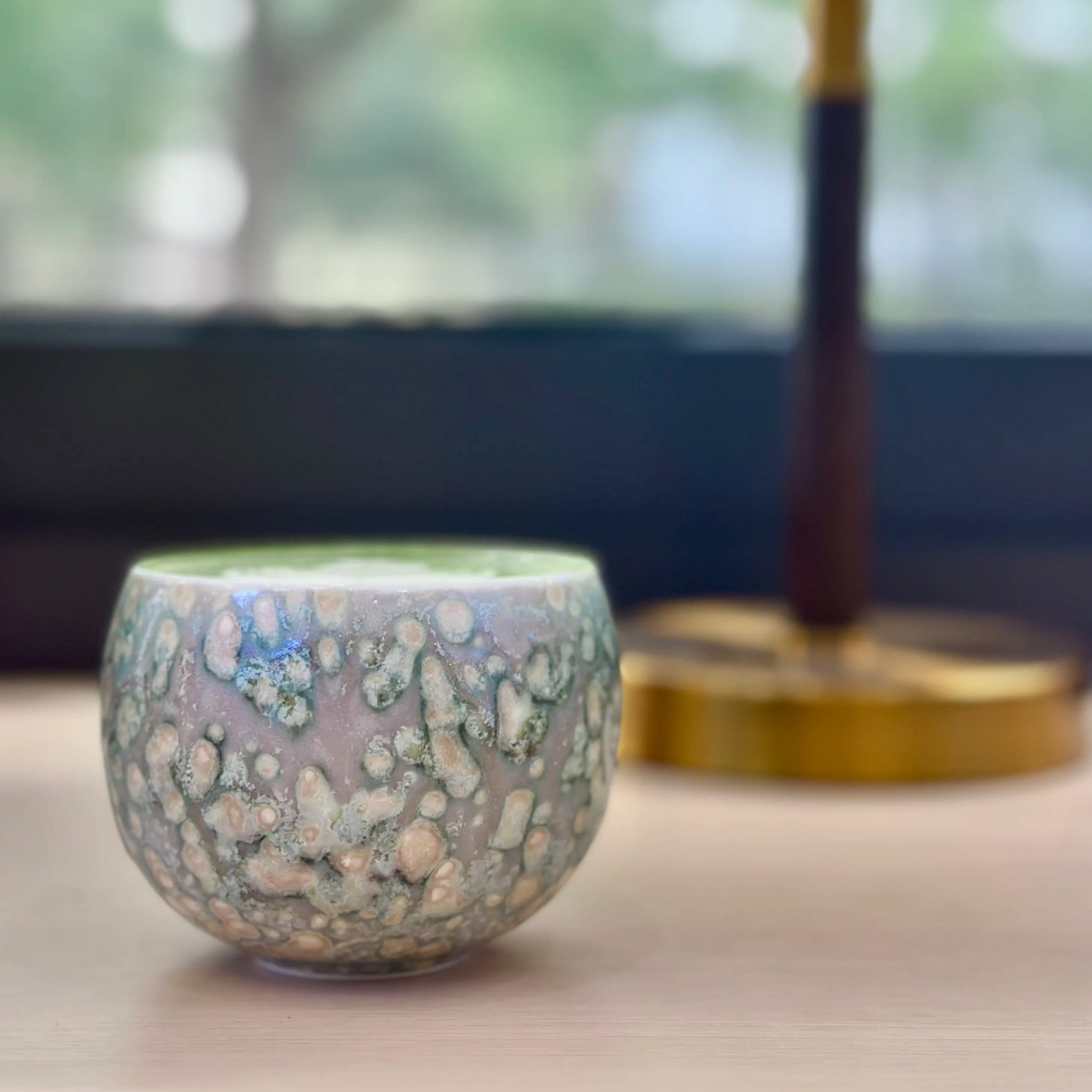 A cozy lifestyle shot of the Glazara Emerald Current matcha latte cup resting on a desk, beautifully illuminated by a backlit window to highlight its vibrant, flowing grey green glaze.