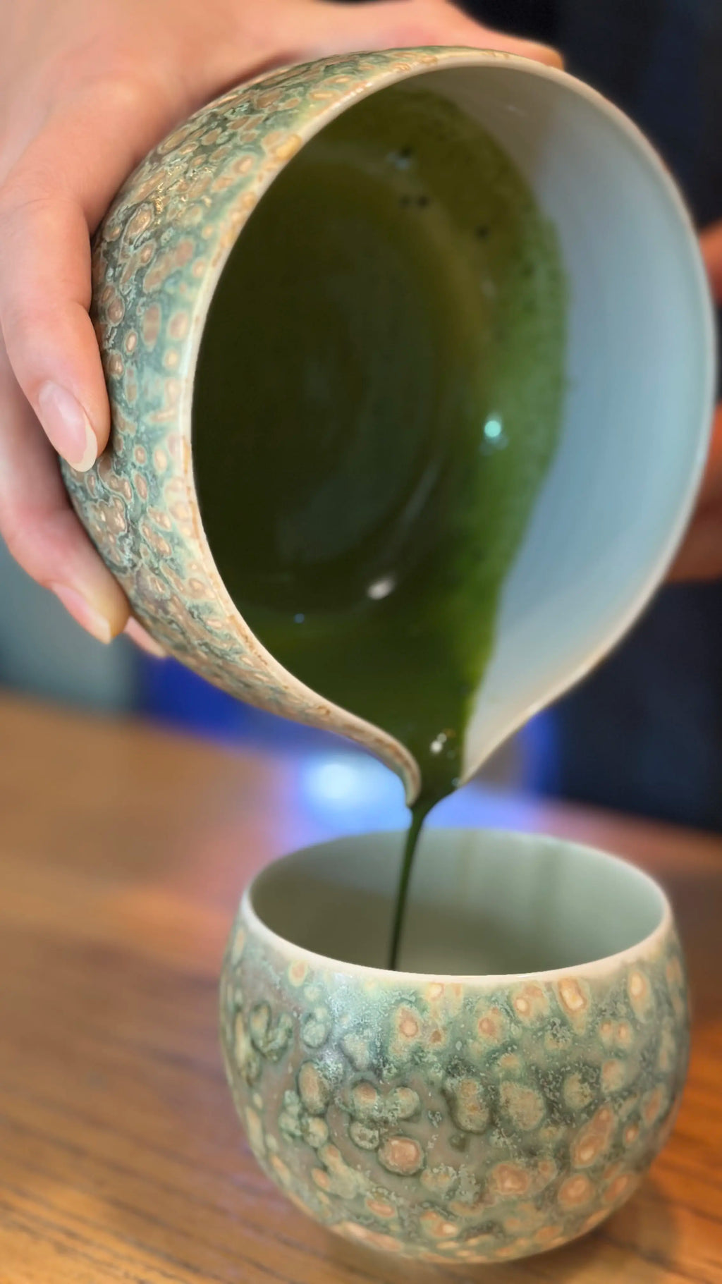 Lifestyle shot of vibrant green tea pouring gracefully from the artisan Glazara Emerald Drift Matcha Bowl into the matching Emerald Drift Matcha Cup.