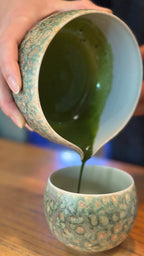 Lifestyle shot of vibrant green tea pouring gracefully from the artisan Glazara Emerald Drift Matcha Bowl into the matching Emerald Drift Matcha Cup.