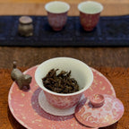 A pink and white porcelain gaiwan sits on a saucer in a tea setting, with loose-leaf tea inside. Two matching cups and a small clay tea pet are blurred in the background on a dark mat.