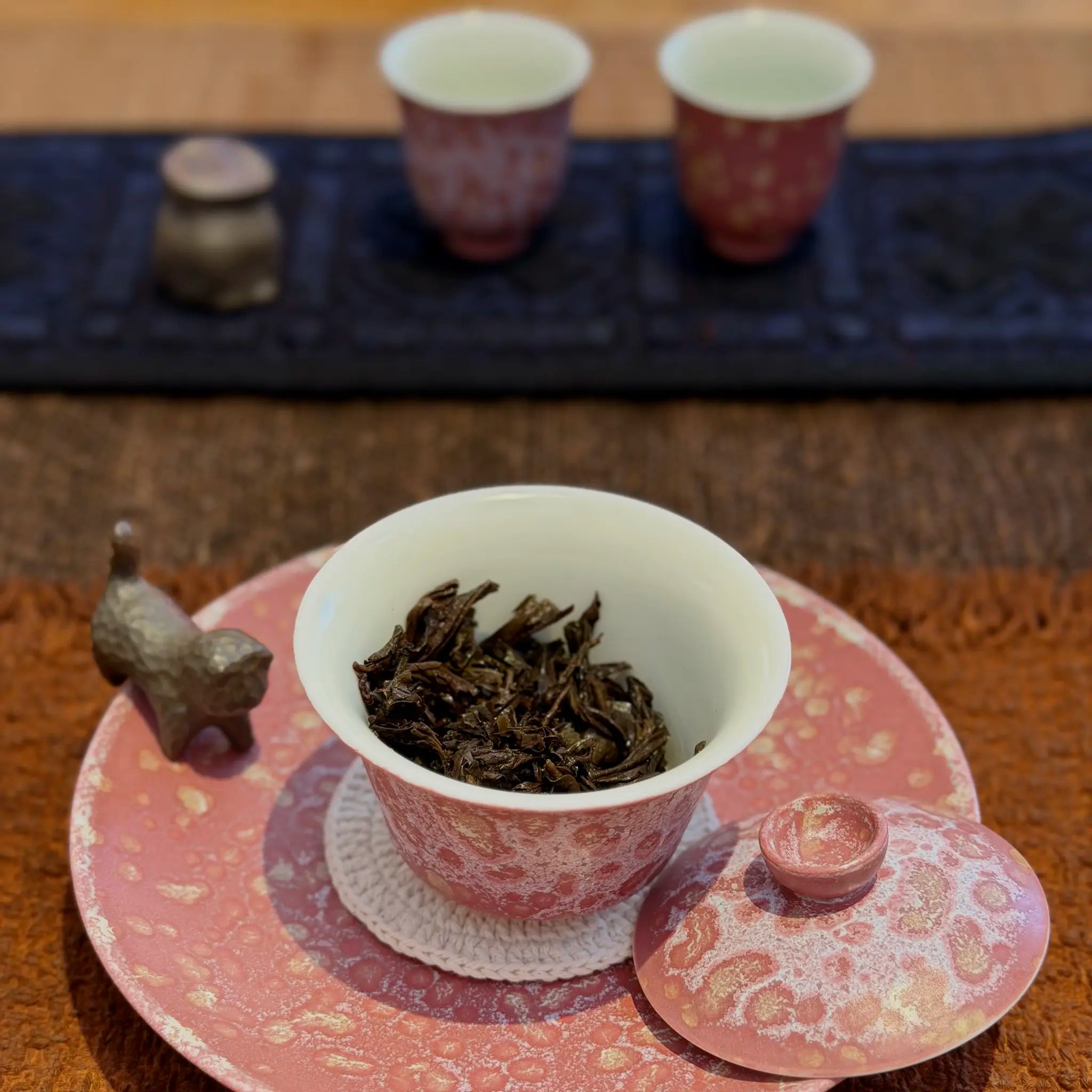 A pink and white porcelain gaiwan sits on a saucer in a tea setting, with loose-leaf tea inside. Two matching cups and a small clay tea pet are blurred in the background on a dark mat.