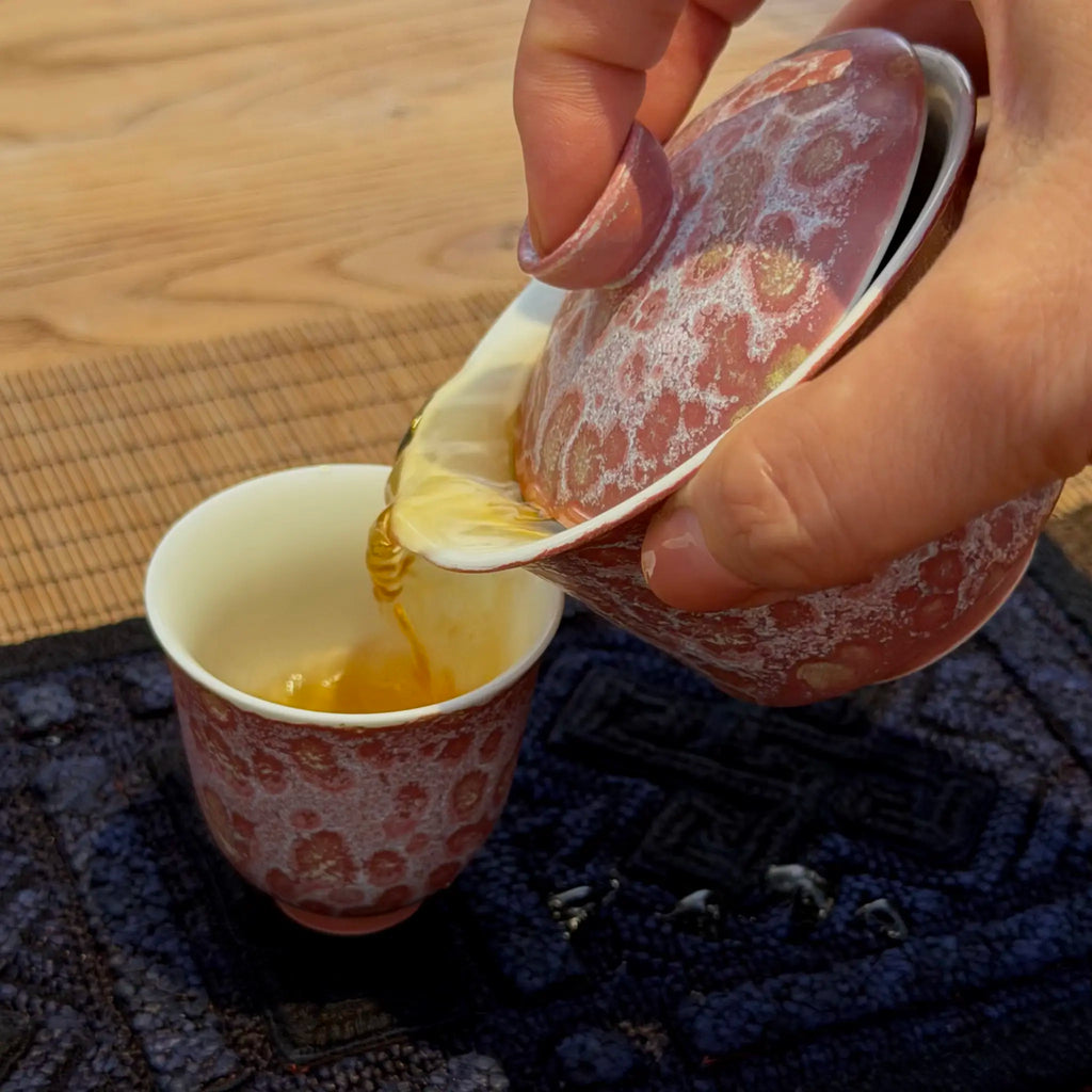 A hand pouring amber tea from a pink and white patterned gaiwan into a matching pink and white patterned cup, set on a dark blue textured mat over a wooden table.