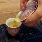 A hand pouring amber tea from a pink and white patterned gaiwan into a matching pink and white patterned cup, set on a dark blue textured mat over a wooden table.