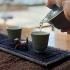 Tea being poured from a gaiwan into a cup on a textured surface.