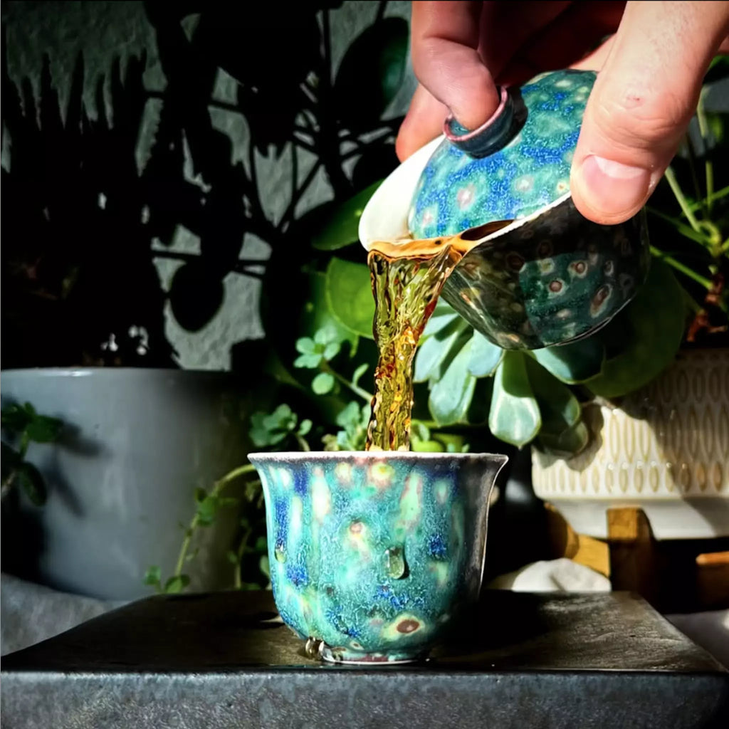 Tea being poured from a pot into a cup with a decorative pattern, set against a natural background.