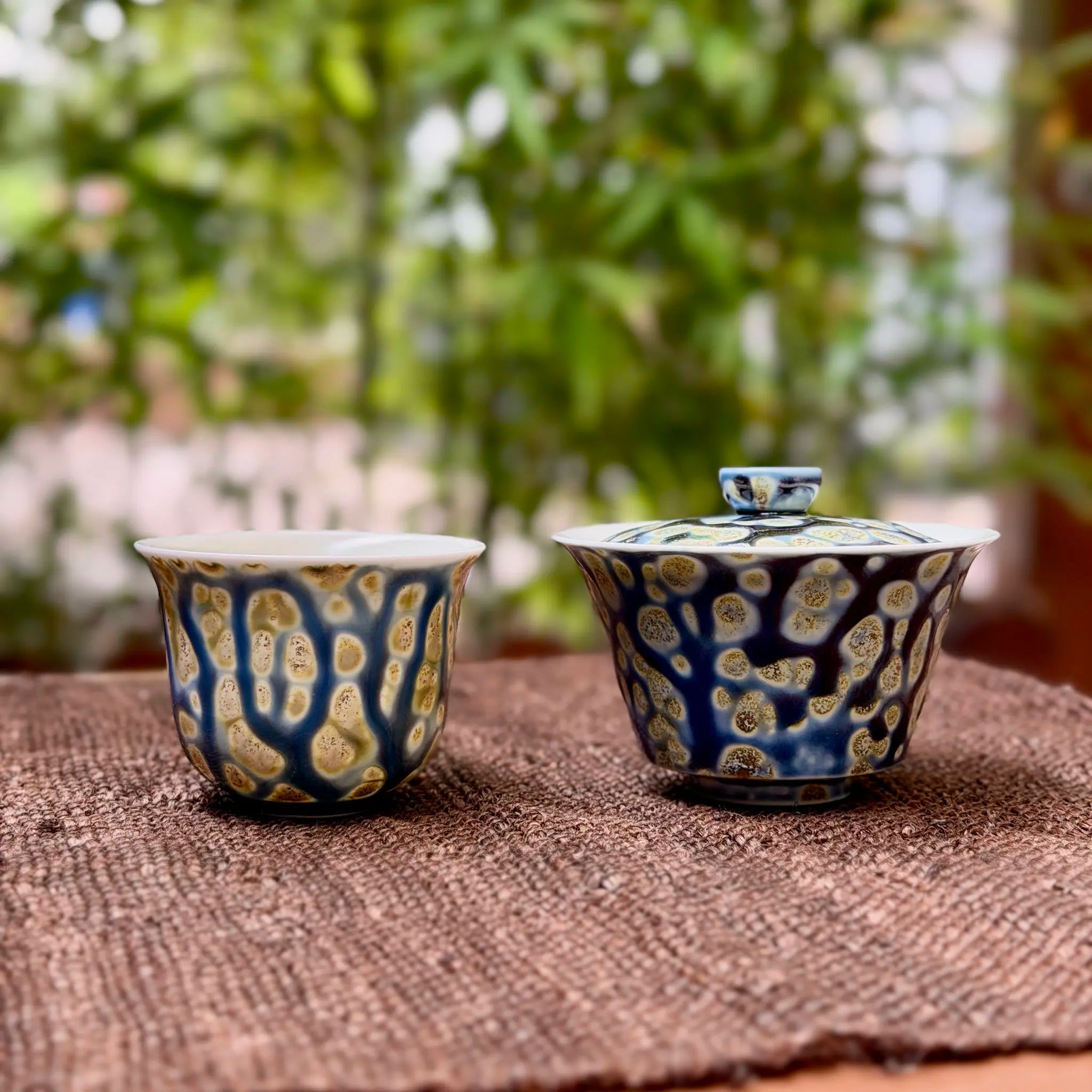 One porcelain gaiwan and one matching teacup with blue and gold patterns on a textured surface with a blurred green background