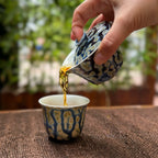 Person pouring tea from a decorative gaiwan into a matching cup on a textured surface with a blurred natural background.