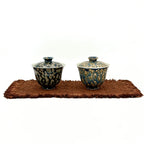 Two porcelain gaiwans with decorative patterns on a brown mat against a white background