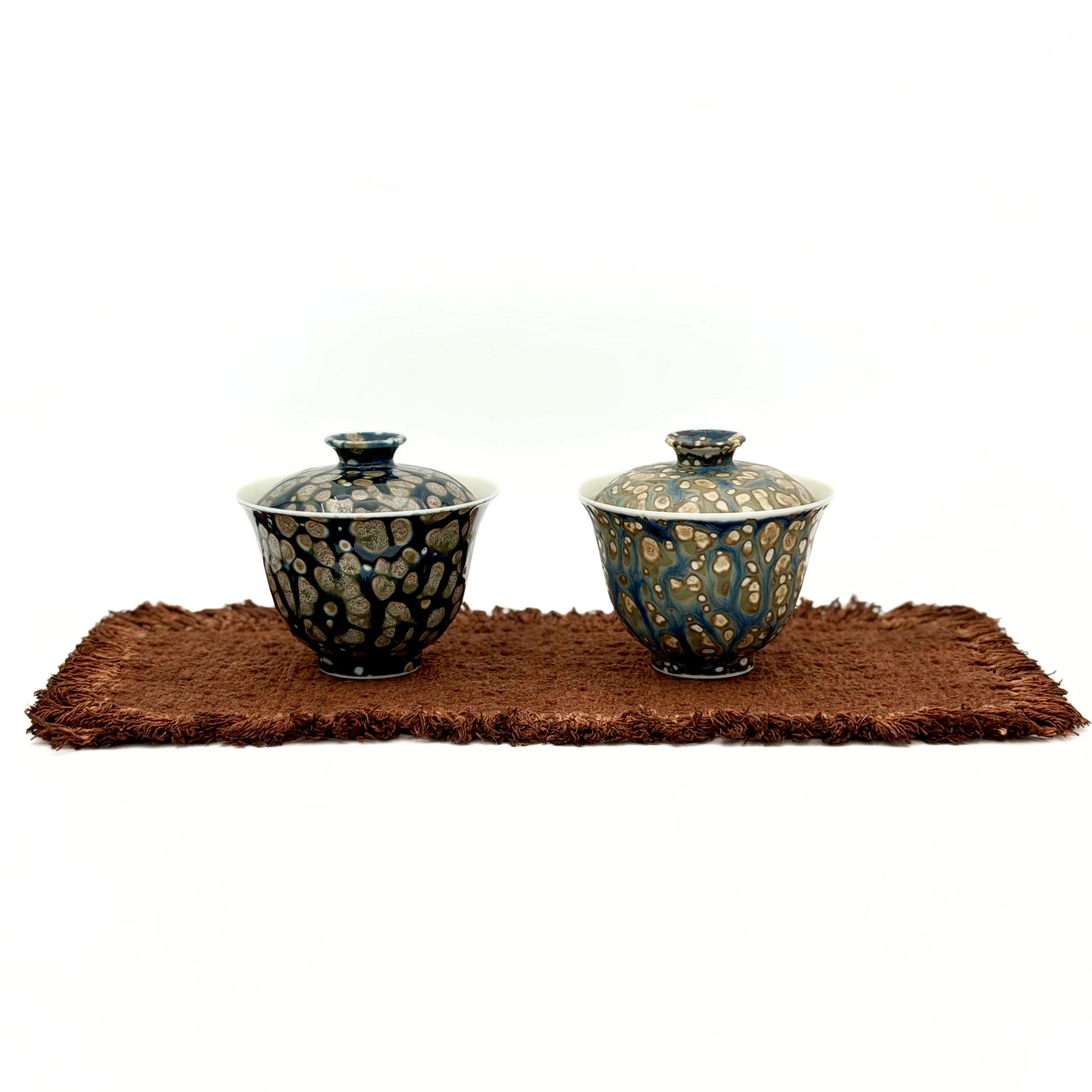 Two porcelain gaiwans with decorative patterns on a brown mat against a white background