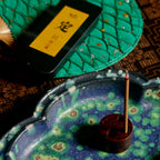 A close-up styled product shot showing a blue and green ceramic incense holder with a single incense stick, alongside a black tablet with a yellow label and a green textured mat on a dark patterned surface.