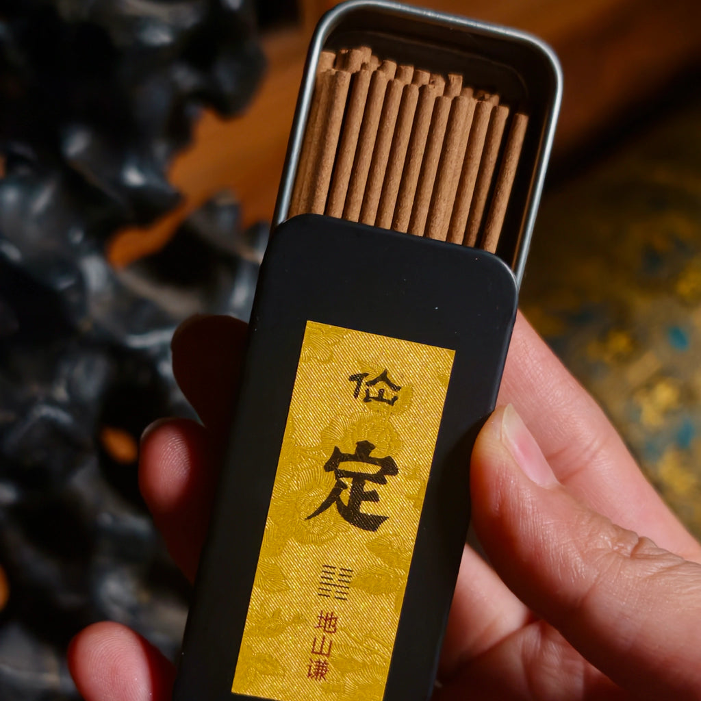 A hand holds an open rectangular metal tin of incense sticks. The tin has a black exterior with a large gold label featuring black Chinese characters, and the incense sticks inside are light brown.