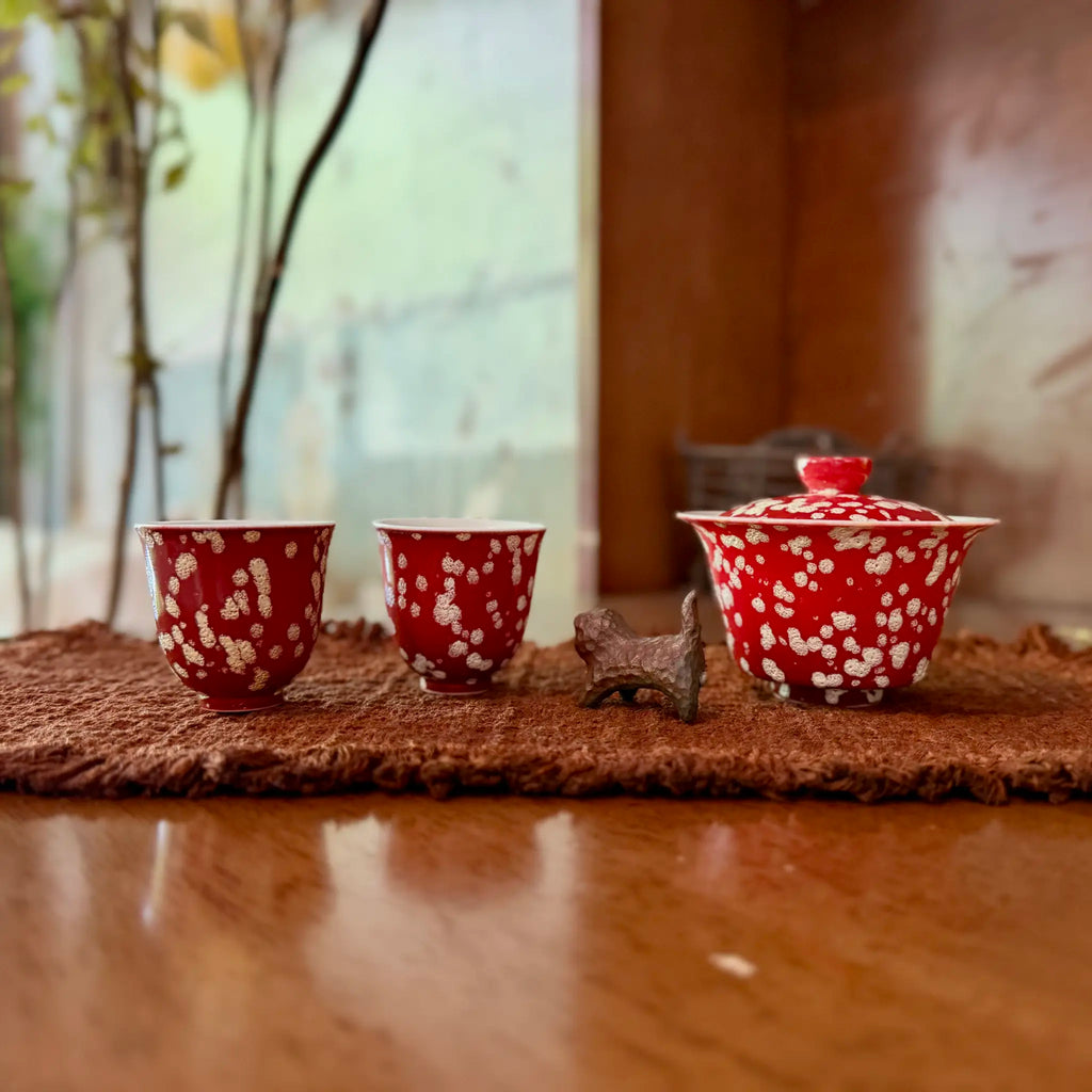 A lifestyle photograph of the Phoenix Spark Chinese Wedding Tea Set, featuring one ceramic gaiwan and two matching tasting teacups resting on a woven tea mat indoors with a softly blurred background.