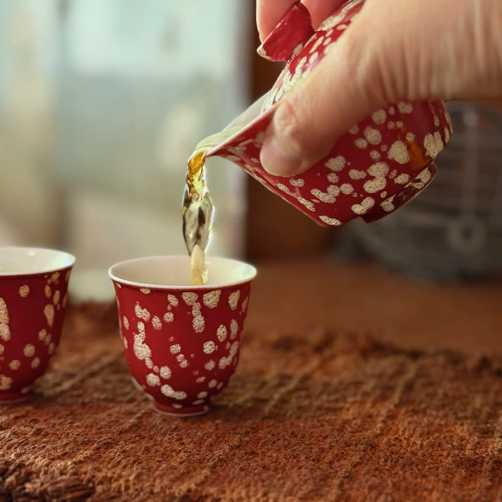 A close-up shot of the Phoenix Spark Chinese wedding gaiwan pouring freshly brewed tea into one of the two matching tasting teacups on a tea mat.