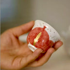 Hand holding a small ceramic teacup with a red and gold design on a blurred background