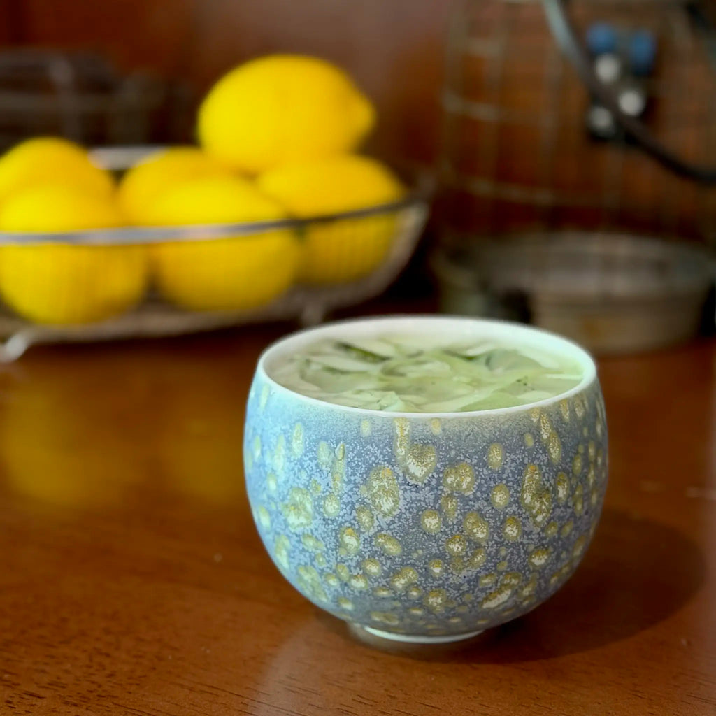 A Glazara Promenade Breeze glass cup filled with a layered iced matcha latte and ice cubes, sitting on a table with a softly blurred bowl of fresh lemons in the background.