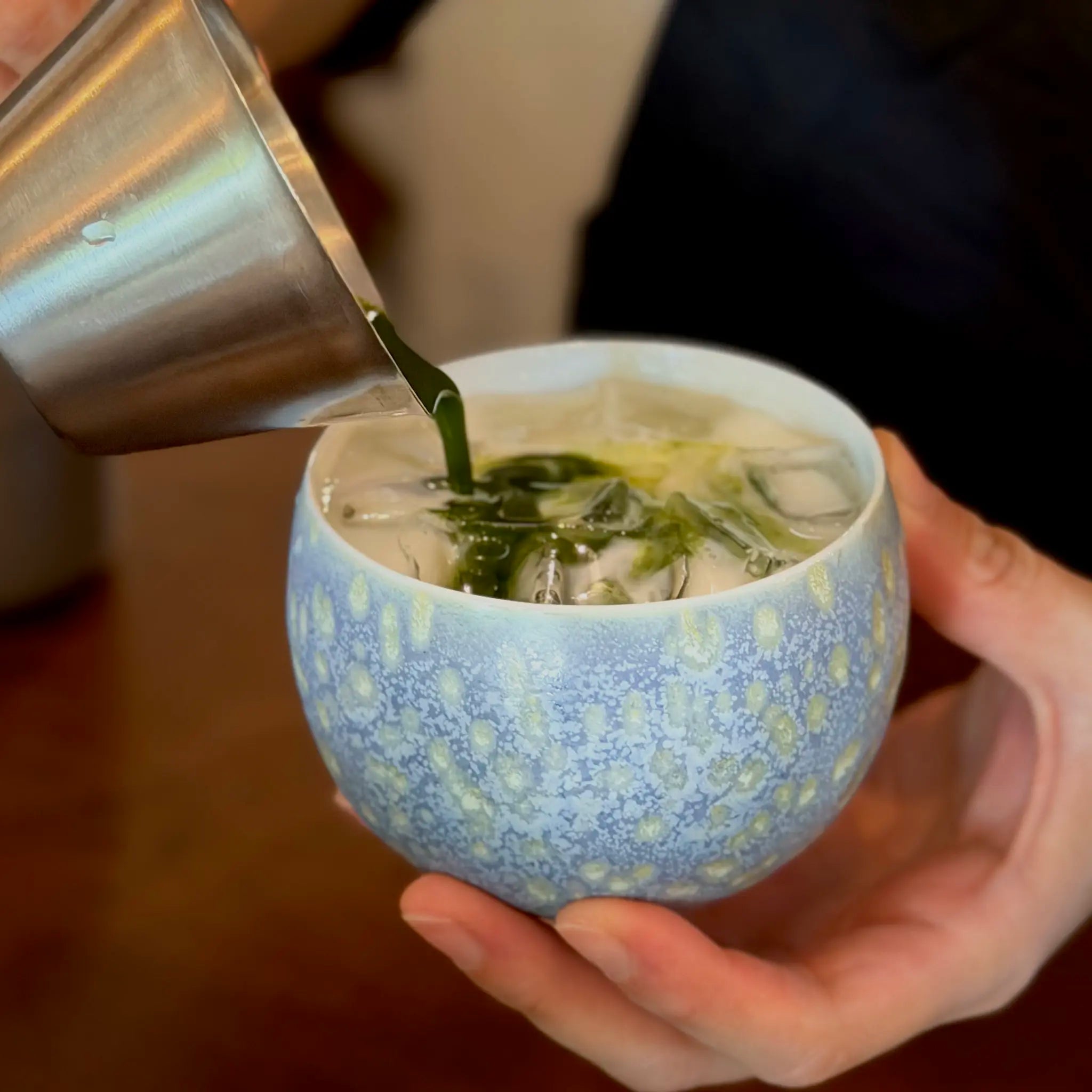 Person pouring green liquid matcha into a blue ceramic cup with impressionism style patterns