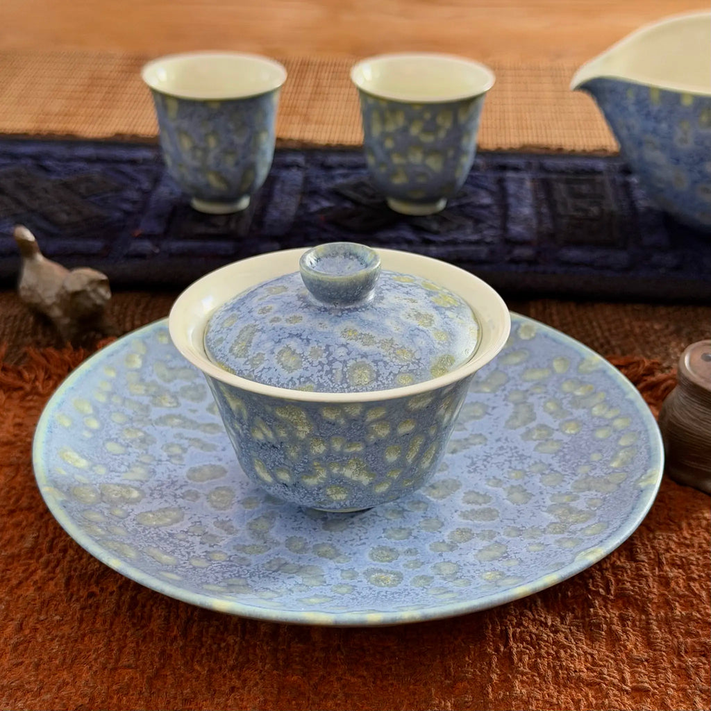 A ceramic tea set with a blue and beige patterned lidded tea bowl on a matching saucer, with two matching cups and a pitcher in the background on a woven mat.