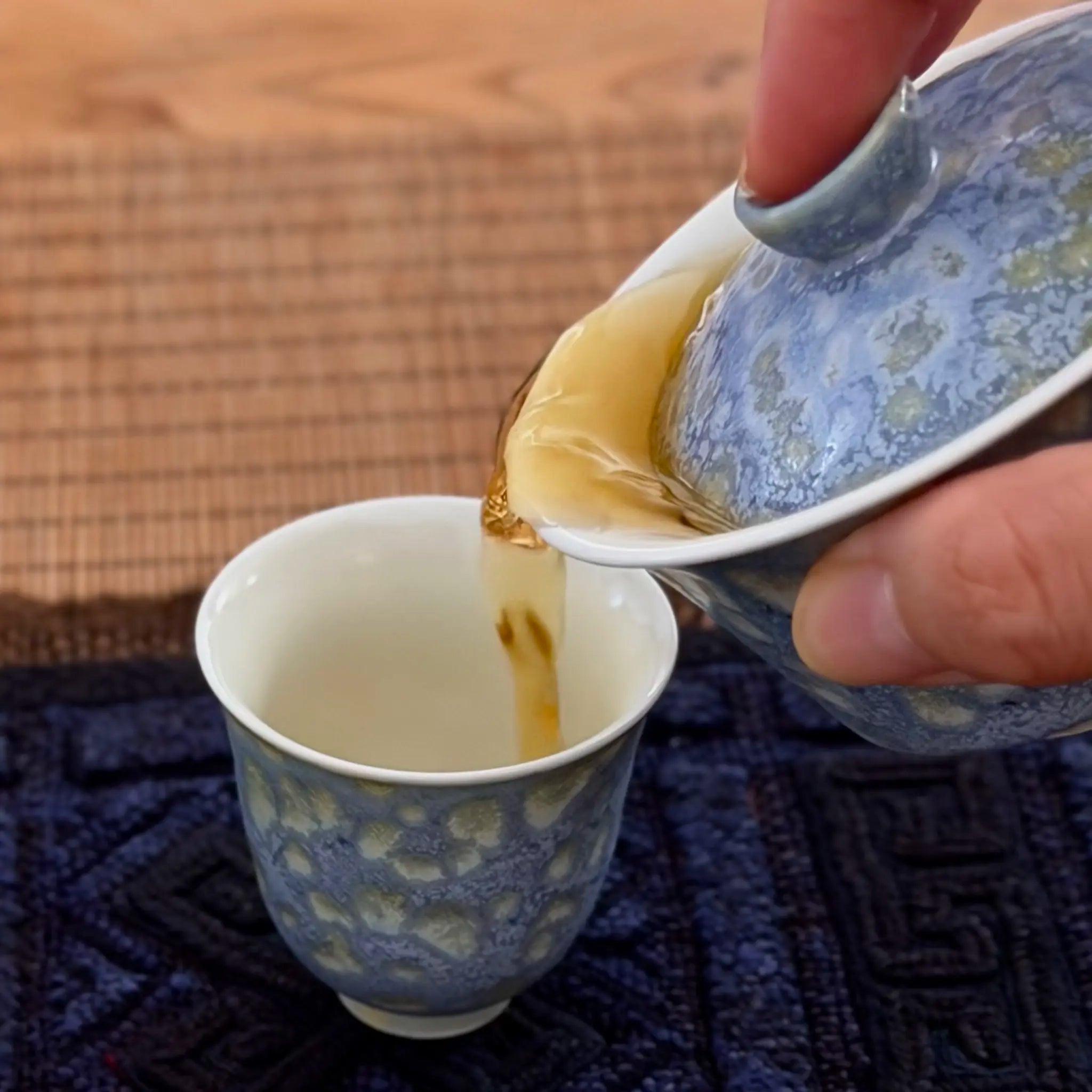 A close-up of a hand pouring tea from a blue and white glazed gaiwan into a matching small tea cup, set on a dark patterned cloth over a woven mat.