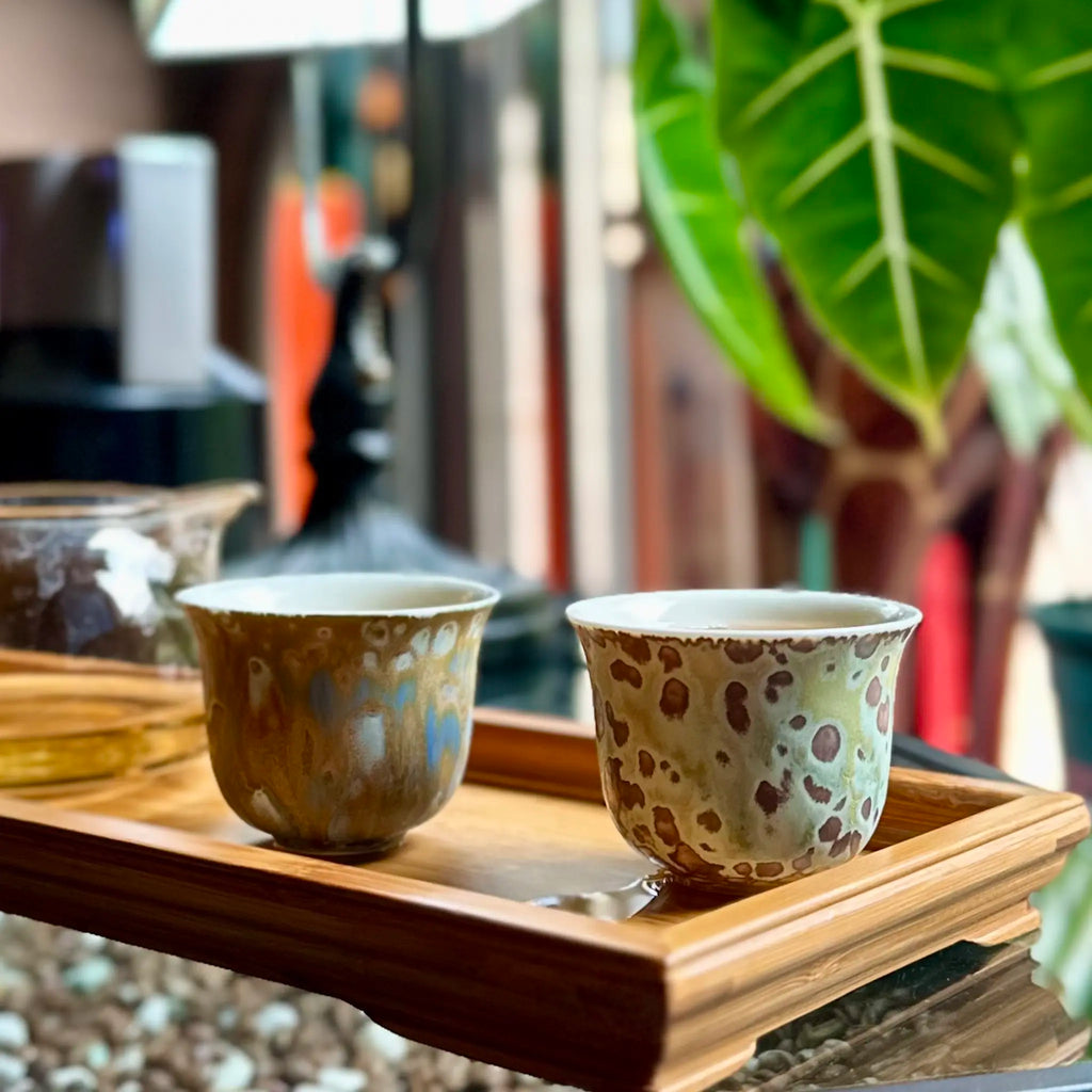 Two porcelain teacups with speckled patterns on a wooden tray, with a blurred indoor background.