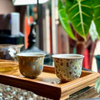 Two porcelain teacups with speckled patterns on a wooden tray, with a blurred indoor background.