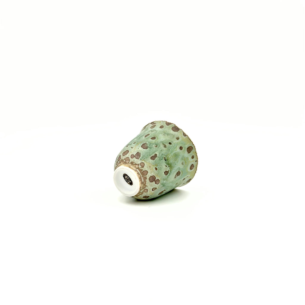 Green ceramic cup with brown speckles on a white background featuring artisan's signature at the bottom.