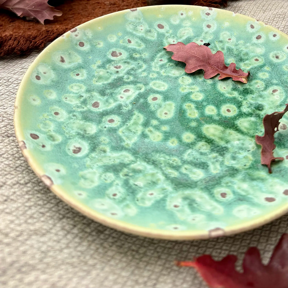 A handmade ceramic plate with a textured, mottled glaze in shades of mint green, turquoise, and cream, decorated with small brown speckles. The plate is styled with dried autumn leaves on a neutral woven fabric background.
