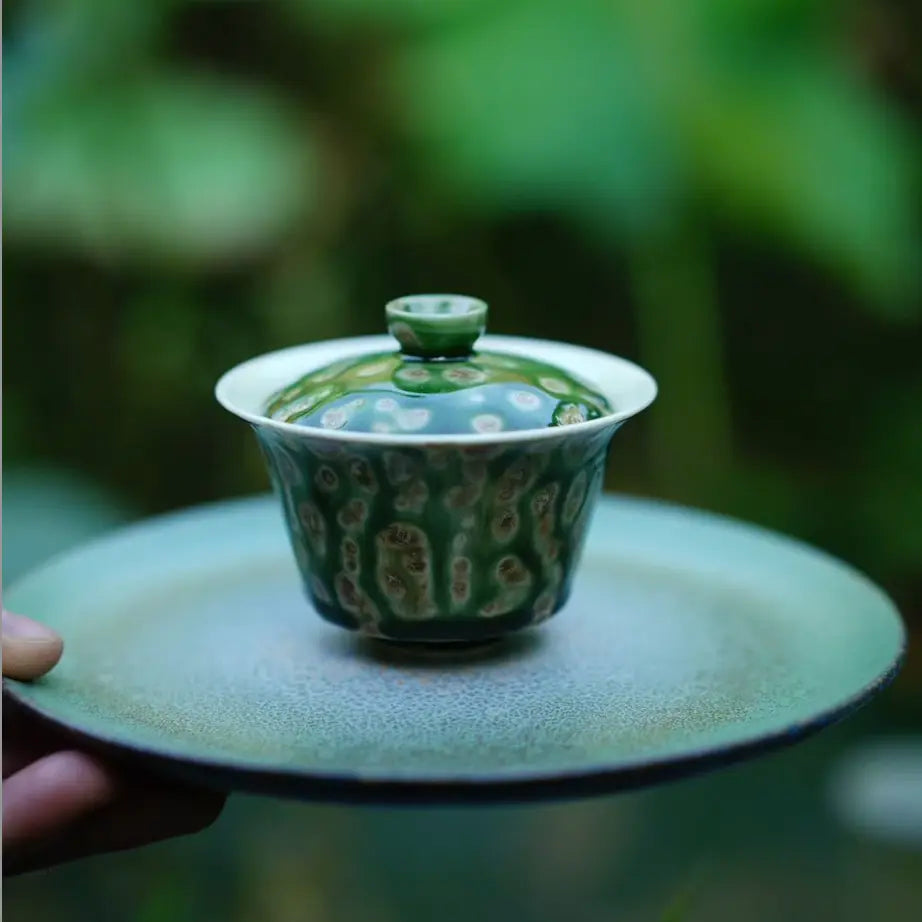 A serene outdoor lifestyle shot holding the Glazara Spring Willow Elemental Gaiwan on a green tea boat against a backdrop of a lush lotus pond, blending traditional teaware with nature.