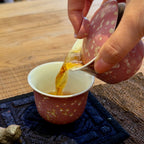 A close-up of amber tea being poured from a pink ceramic gaiwan with gold speckles into a matching pink ceramic cup with a white interior, set on a dark woven tea mat over a wooden table.