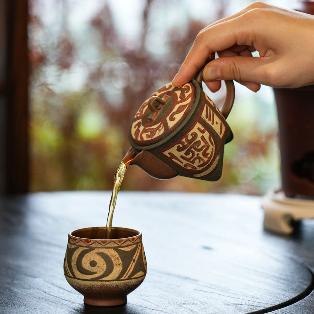 Amber tea gracefully pouring from the spout of the Cloud Thunder Yixing teapot into a matching white-slip painted teacup during a Gongfu tea session.