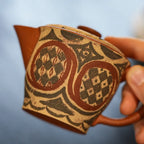Close-up of a hand gently holding the Ivory Canvas Yixing teapot by the handle, highlighting the intricate details of the hand-painted pottery design.