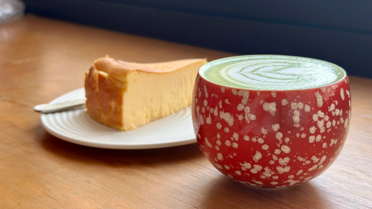 Red ceramic cup with floral pattern containing a green beverage matcha latte on a wooden table next to a piece of cheese cake.