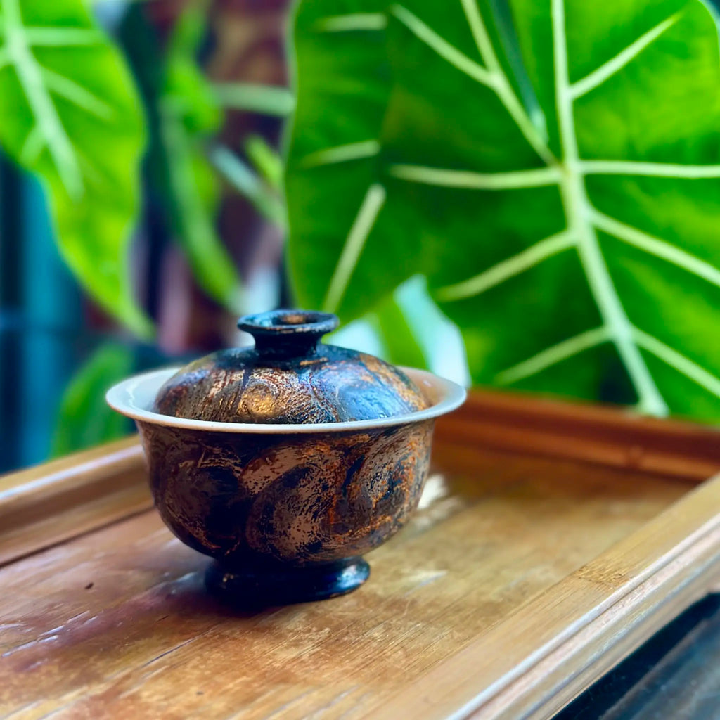 Midnight solo gaiwan styled on a bamboo tea tray against a green indoor plant background.