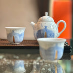 Ceramic teapot and cups with blue and white design on a reflective surface.