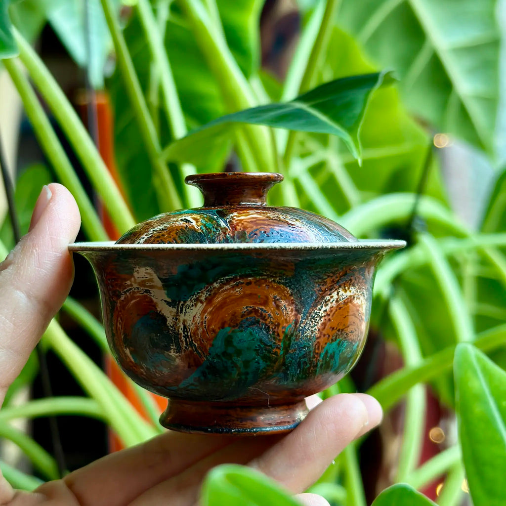 Hand holding an urushi gaiwan against a green plant background